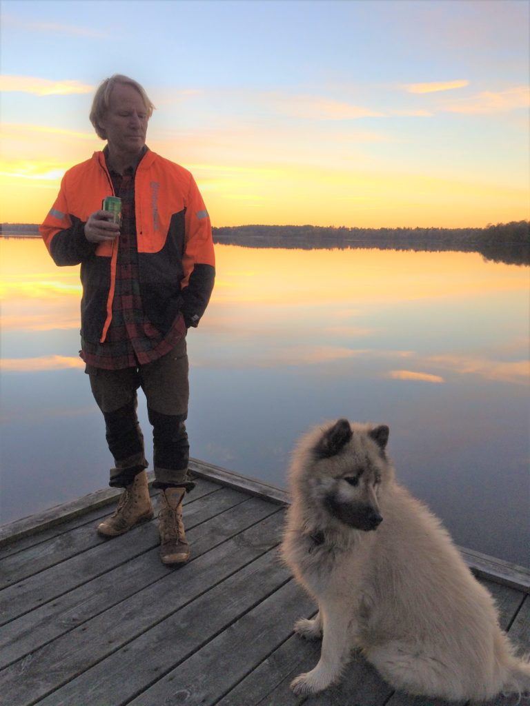 Bengt Söderberg står på en brygga vid en sjö i solnedgång. Bengt är klädd i orange jacka, rödrutig skjorta, beigea byxor och beigea skor. Till höger om Bengt på bryggan sitter en vit hund.