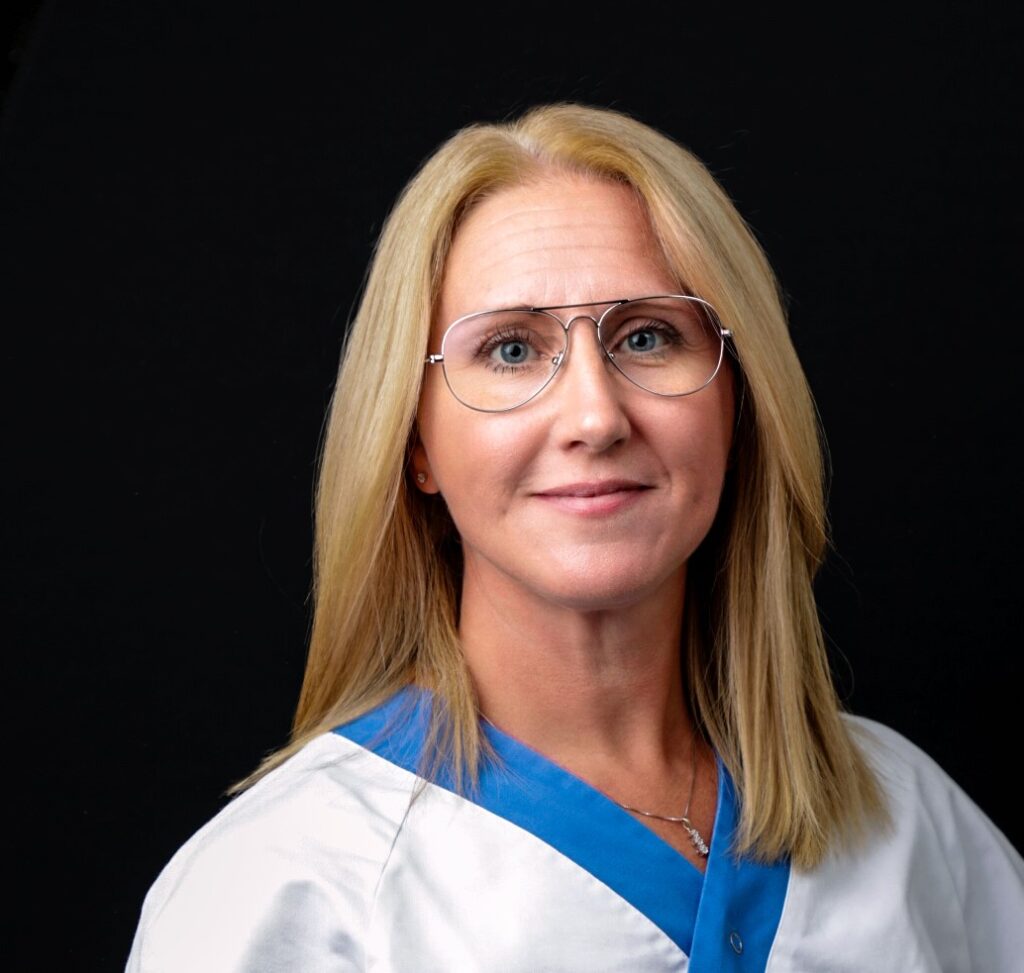 Porträttfoto av Åsa Okhiria. Åsa är klädd i vit läkarrock med blå krage. Åsa har långt, blont hår och glasögon.