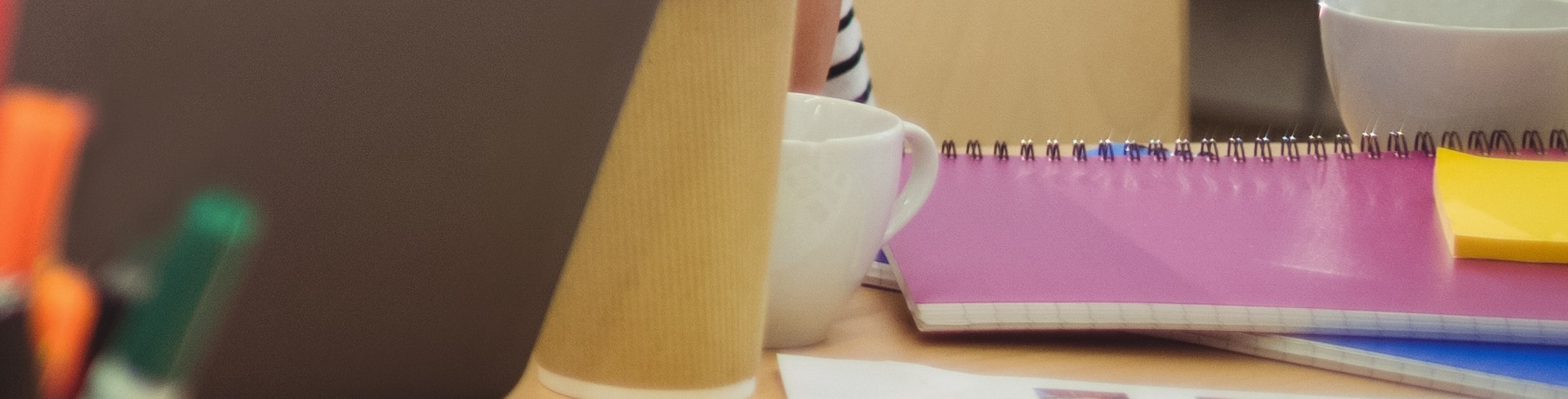 Skrivbord med svart laptop, vit kaffekopp, gula Post-It-lappar och ett par anteckningsböker, rosa respektive blå.