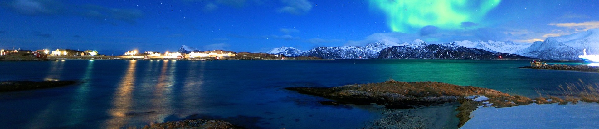 Vinterkall kväll med norrsken över insjö. I horisonten syns en fiskeby till vänster och en snötäckt bergskedja till höger..
